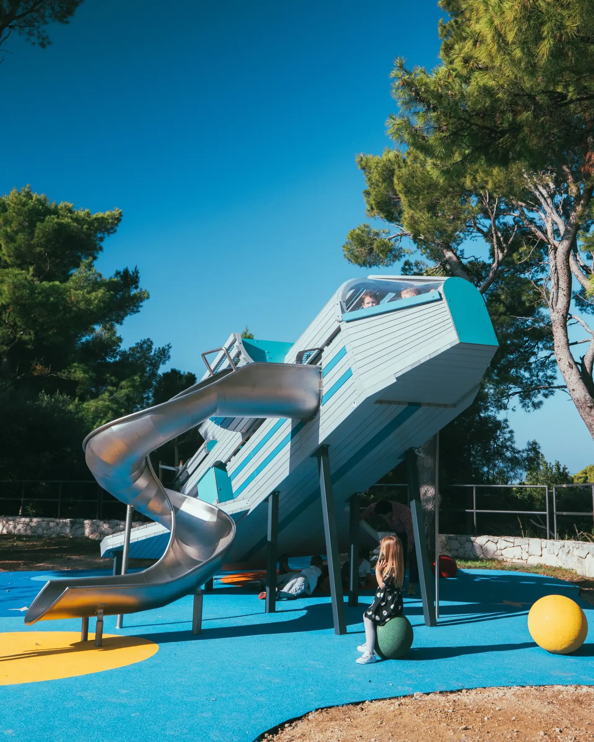 The rocket-shaped playground structure at Makarska Observatory, seen from the side, with its spiral slide and children playing on the surfacing below.