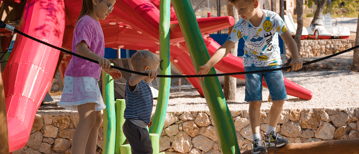 Three children balancing on a rope handrail on a red-and-green, sea-creature-themed play structure at an outdoor playground, with a stone wall in the background.