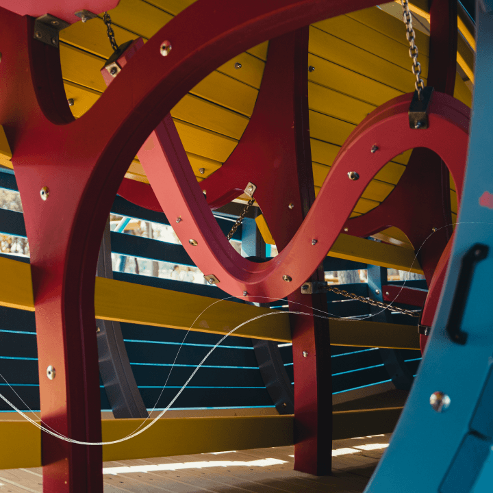 Close-up of a themed playground’s curved red timber structure, with a yellow ceiling, dark teal slats and chain details.