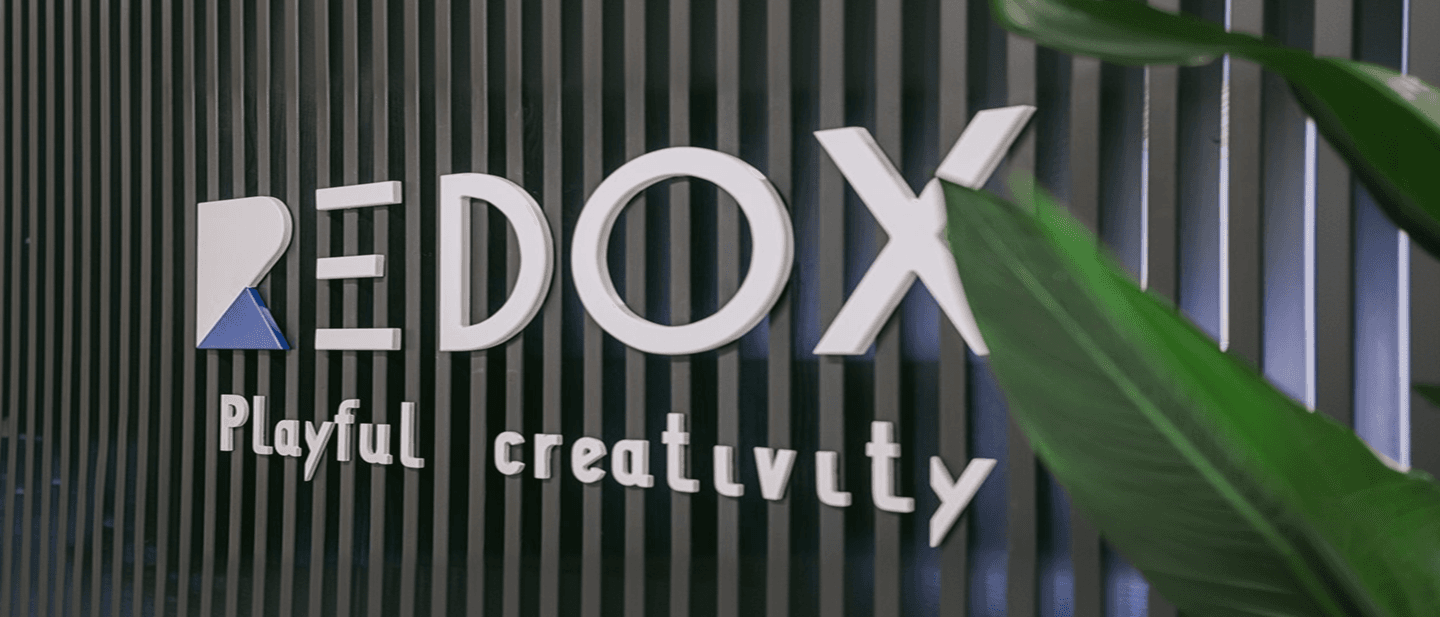 REDOX 3D logo signage reading “Playful creativity”, mounted on a slatted wood panel with a green plant leaf in the foreground.